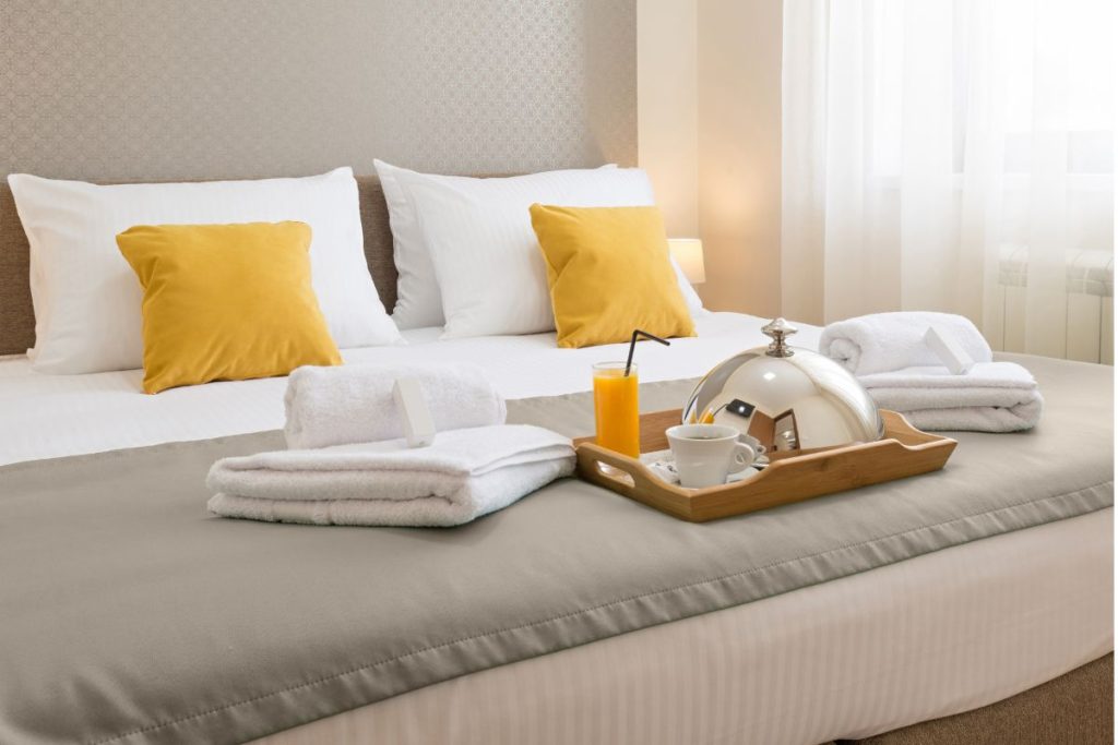 Perfectly made bed with crisp white sheets, yellow accent pillows, folded towels, and a breakfast tray with coffee and juice