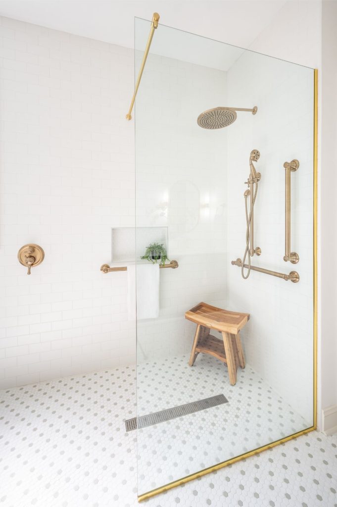 Curbless walk-in shower with brass fixtures, grab bars, teak bench, and white tile in a Chapel HIll bathroom remodel