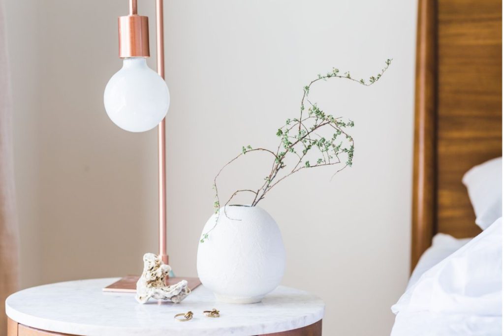 Bedside table with marble top, rose gold lamp, white vase with greenery, and natural decorative objects in a softly lit bedroom