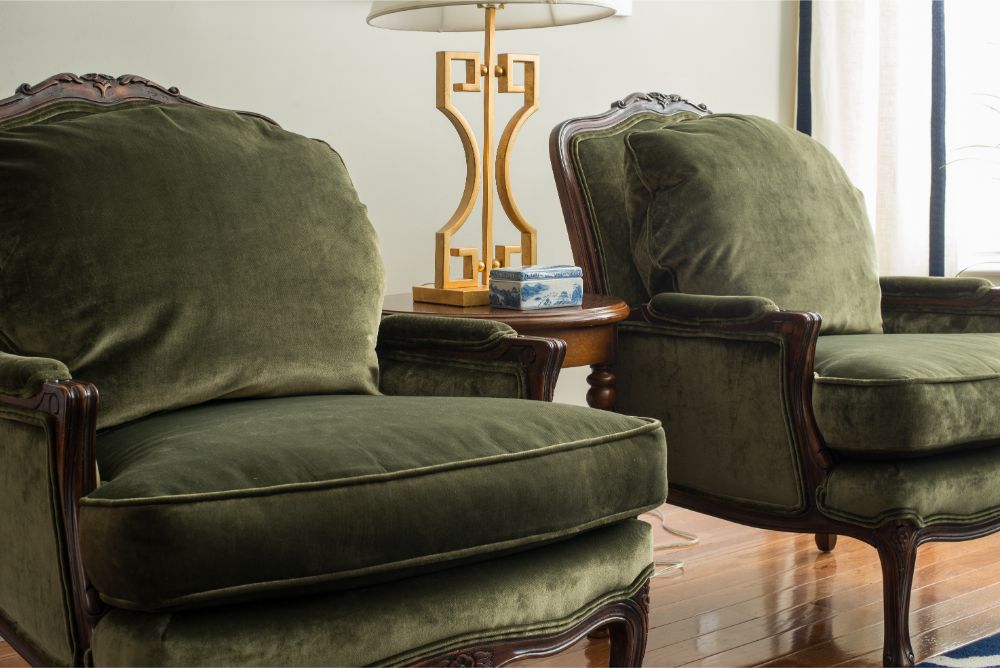 Two olive green velvet armchairs with carved wood frames beside a lamp and side table in a bright living room