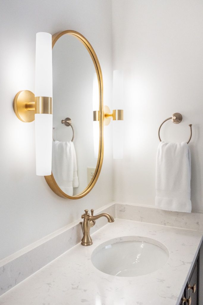 Oval mirror with gold frame and wall sconces above a white marble vanity in a remodeled bathroom in Chapel Hill, North Carolina