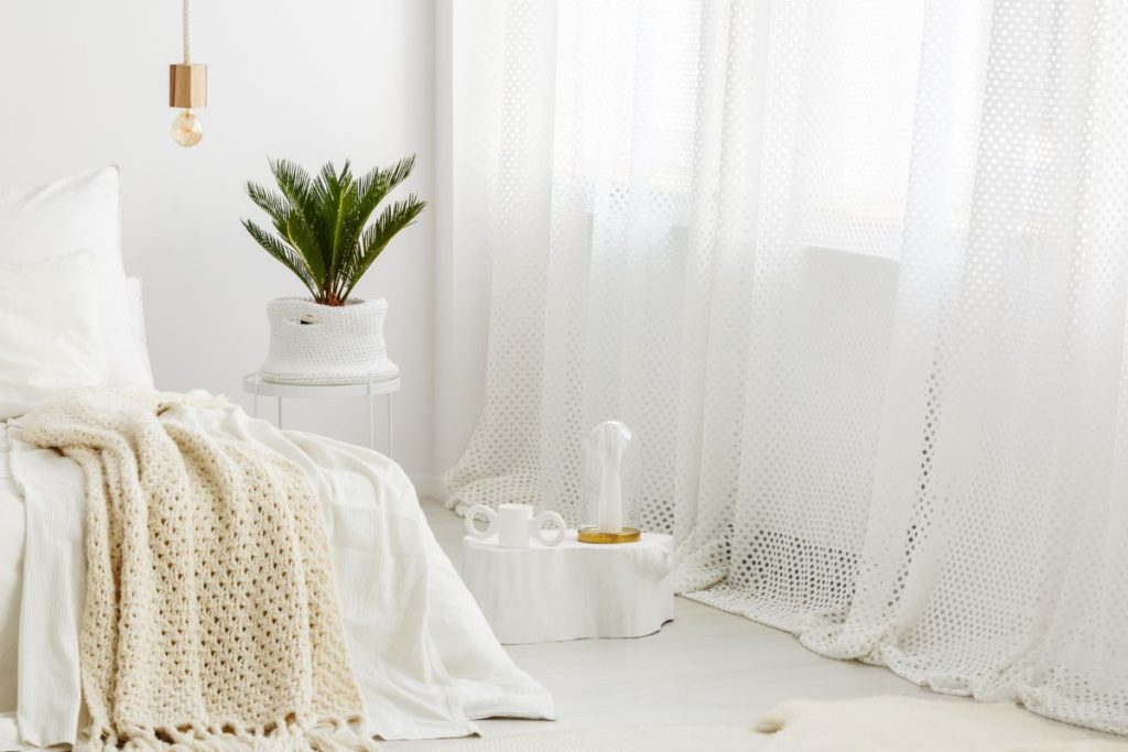 Minimalist white bedroom with sheer textured curtains, knit throw, woven basket planter, and ceramic accessories